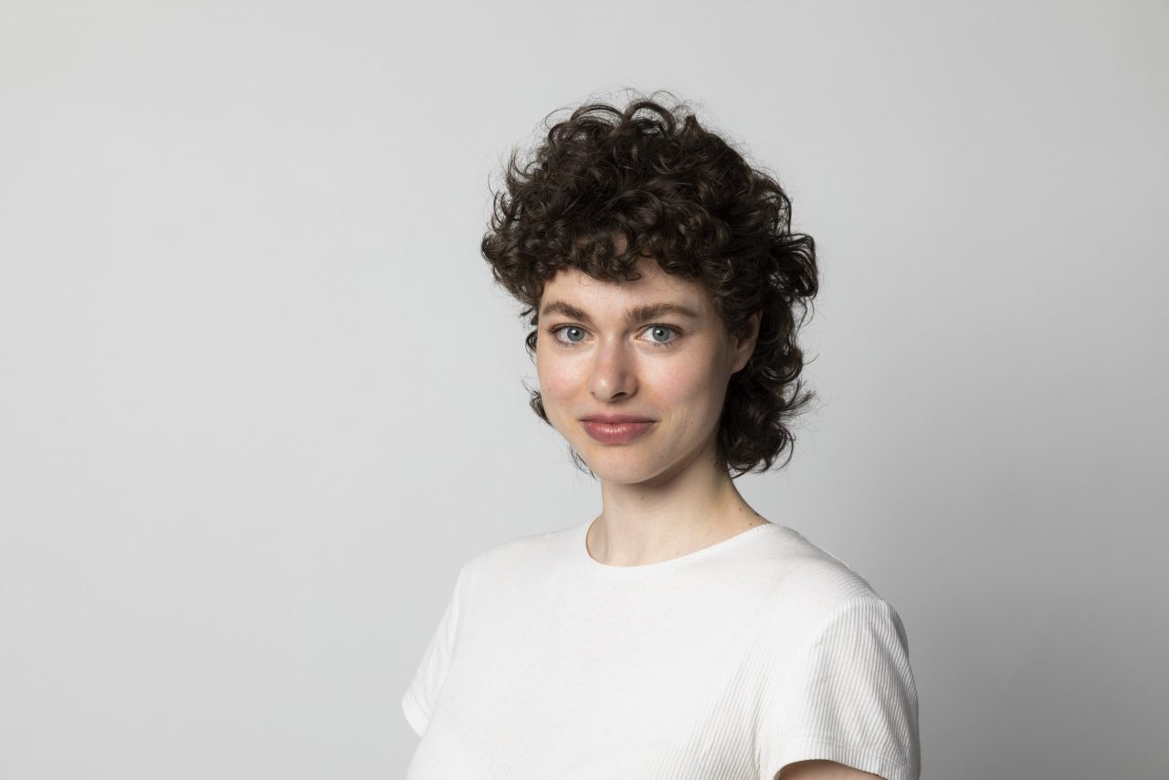 Portrait en couleur sur fond gris clair d'une femme brune souriante aux cheveux bouclés qui porte un T-Shirt blanc