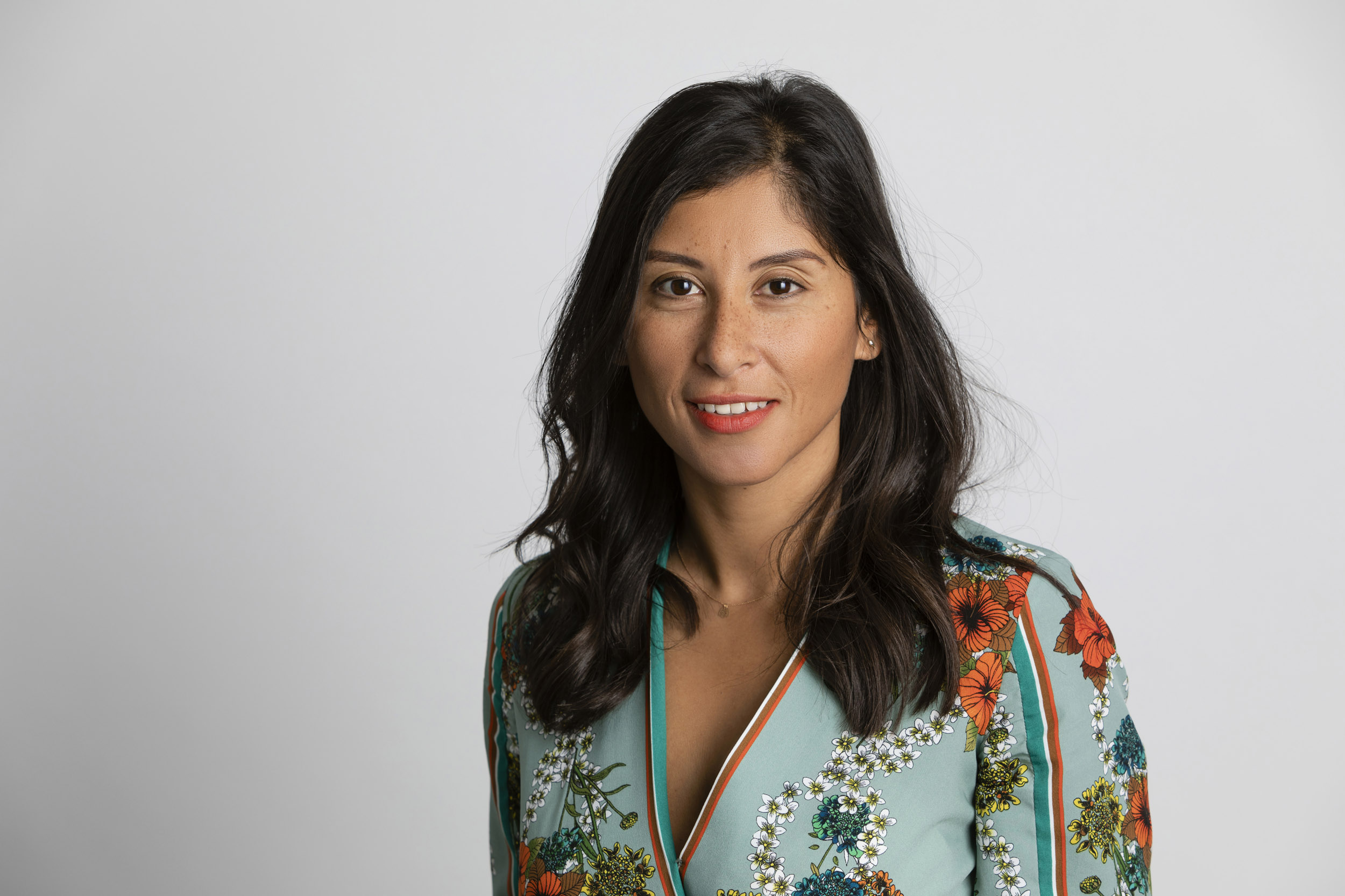 Portrait corporate en studio sur fond clair d'une femme brune souriante aux cheveux longs cadré en plan buste. Elle porte un chemisier à fleurs