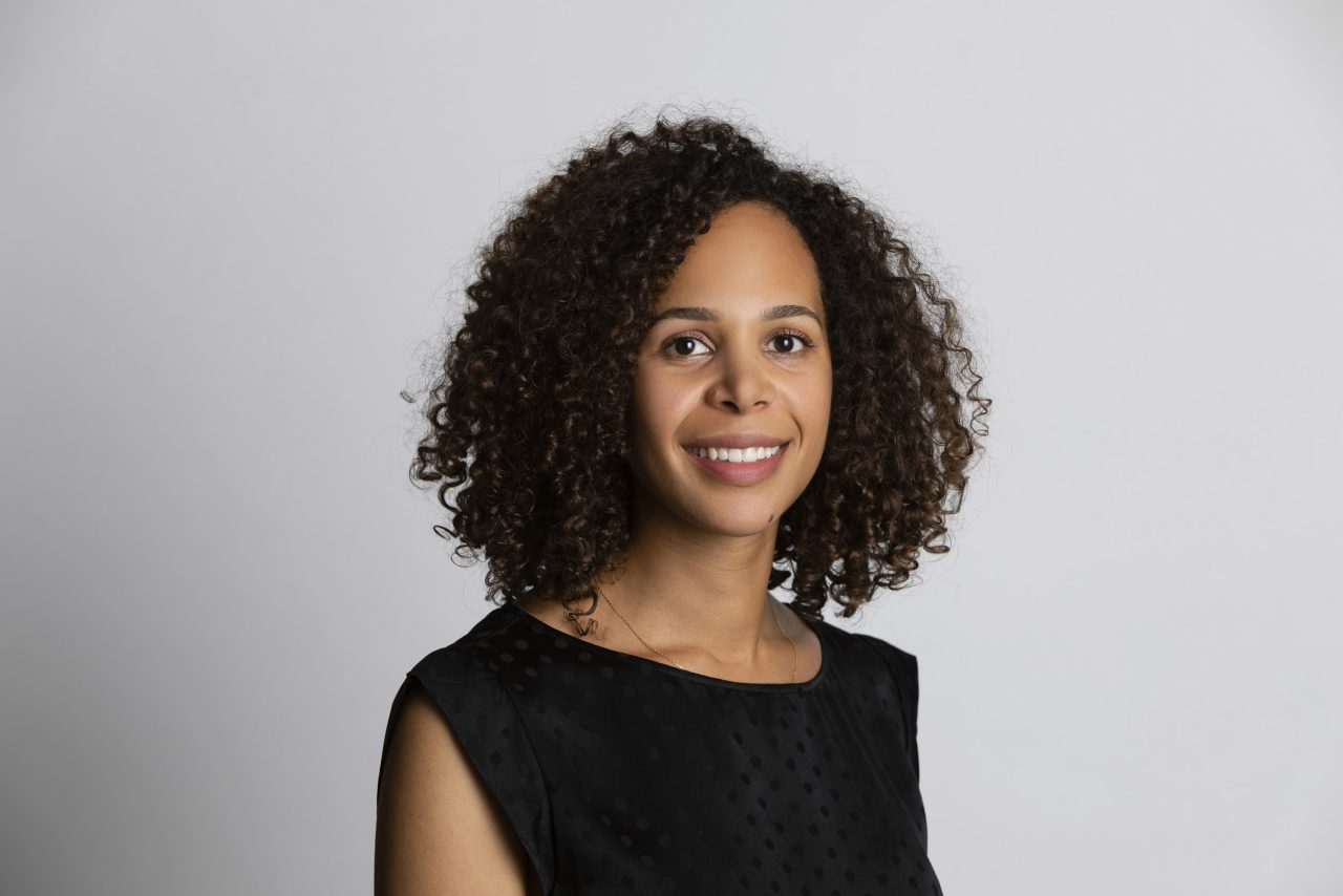 Portrait corporate en studio sur fond clair d'une femme souriante brune cadré en plan buste. Elle porte une robe noir et des cheveux bouclés