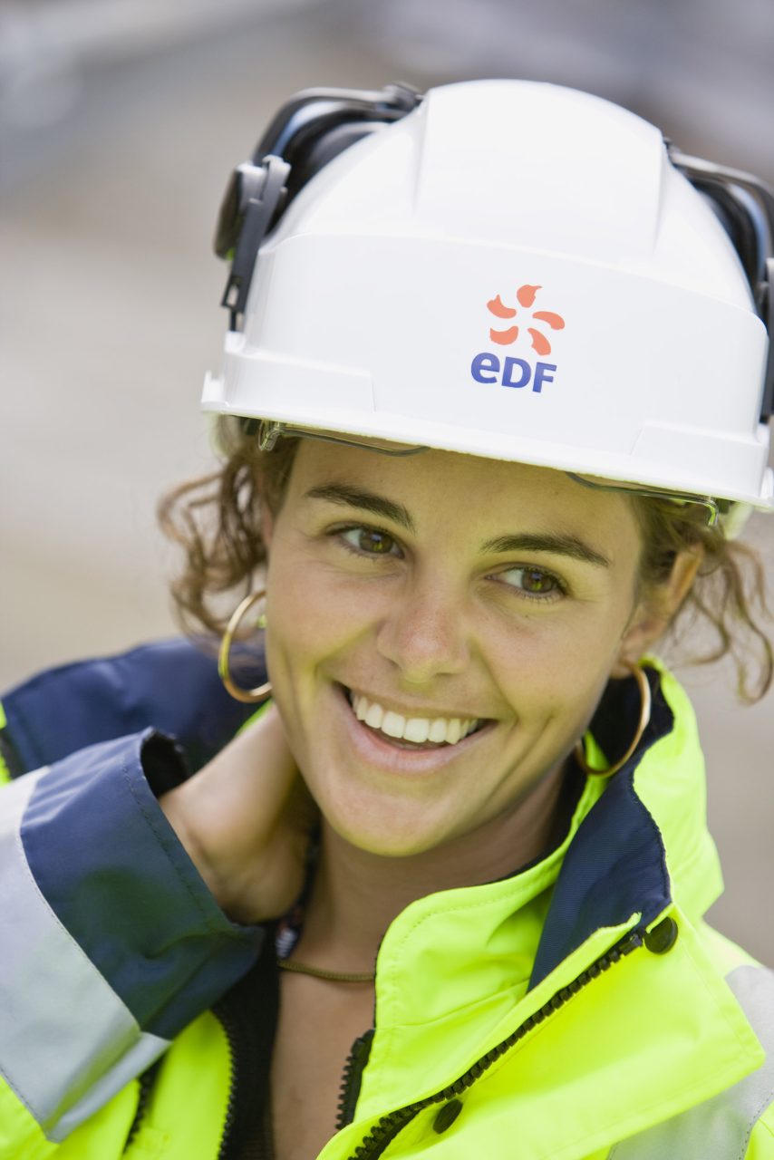 Portrait corporate d'une jeune femme souriante aux yeux bruns, elle porte un casque de chantier avec le logo EDF