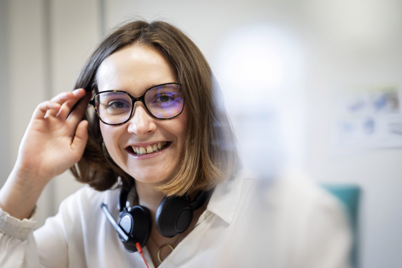 Portrait métier d'une jeune femme souriante assise à son bureau, elle regarde dans l'objectif en remettant en place une mèche de cheveux