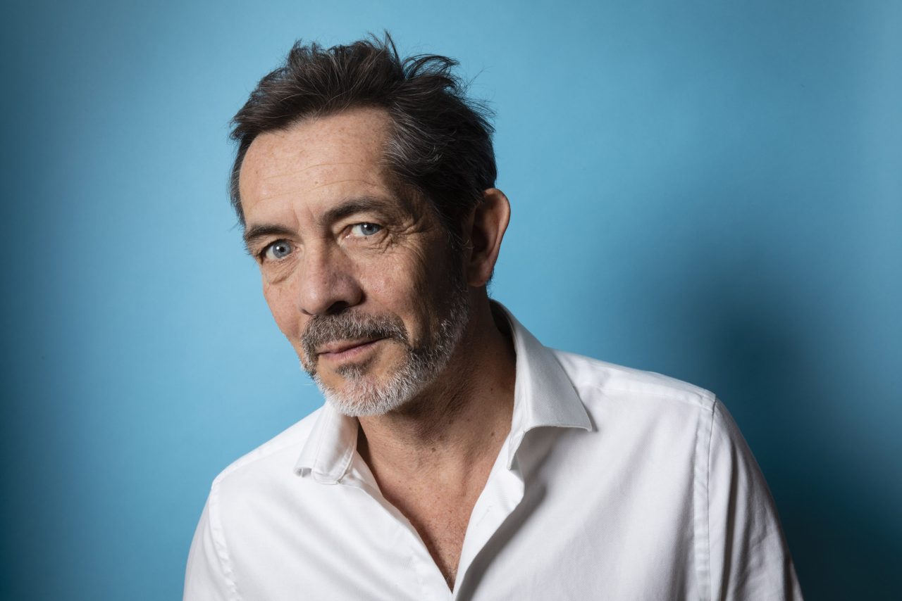 Portrait corporate d'un quinquagénaire brun aux yeux bleus avec une chemise blanche qui pose devant un fond bleu turquoise