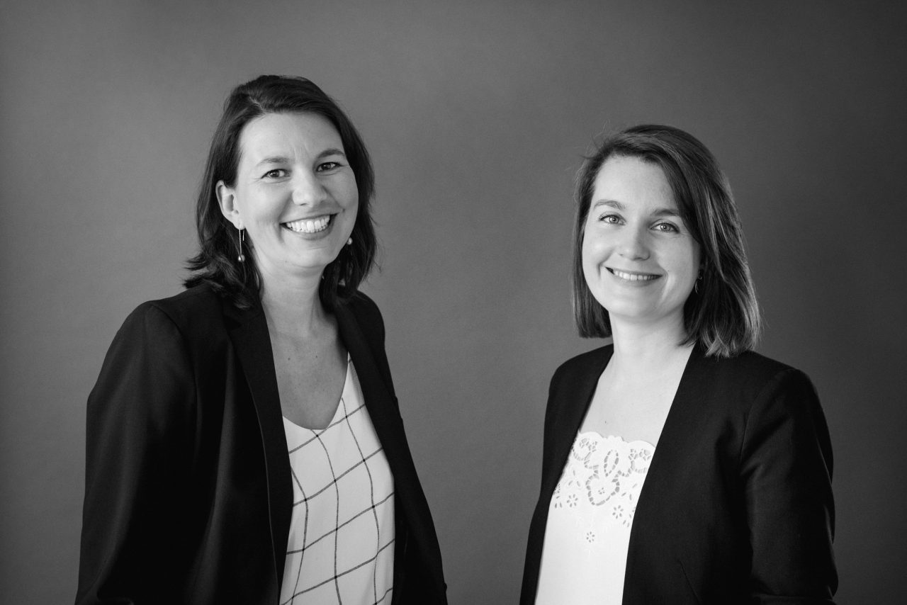 Portrait noir et blanc en duo de 2 femmes avocates souriantes côtes à côtes en plan buste sur fond gris