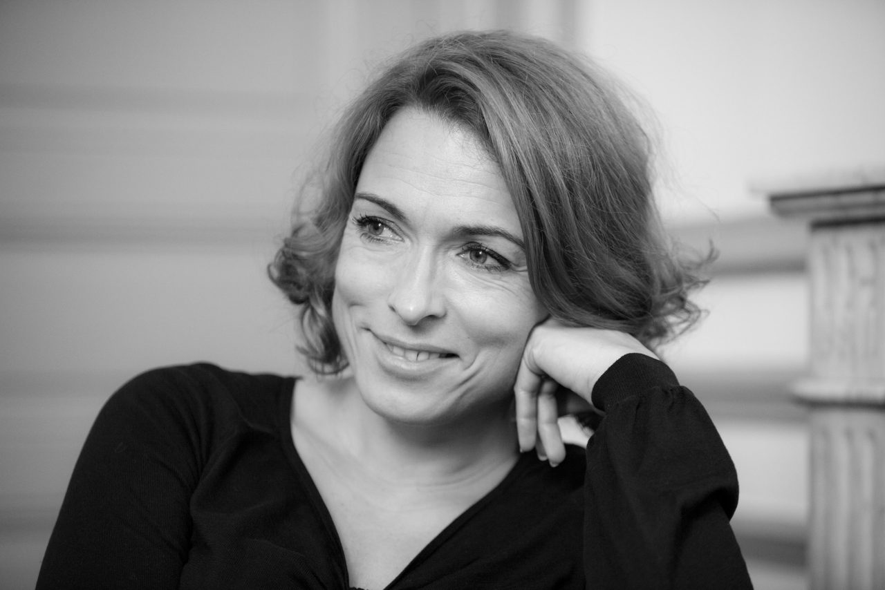 Portrait corporate en noir et blanc d'une dirigeante d'entreprise souriante en réunion dans un salon haussmannien. Elle regarde son interlocuteur