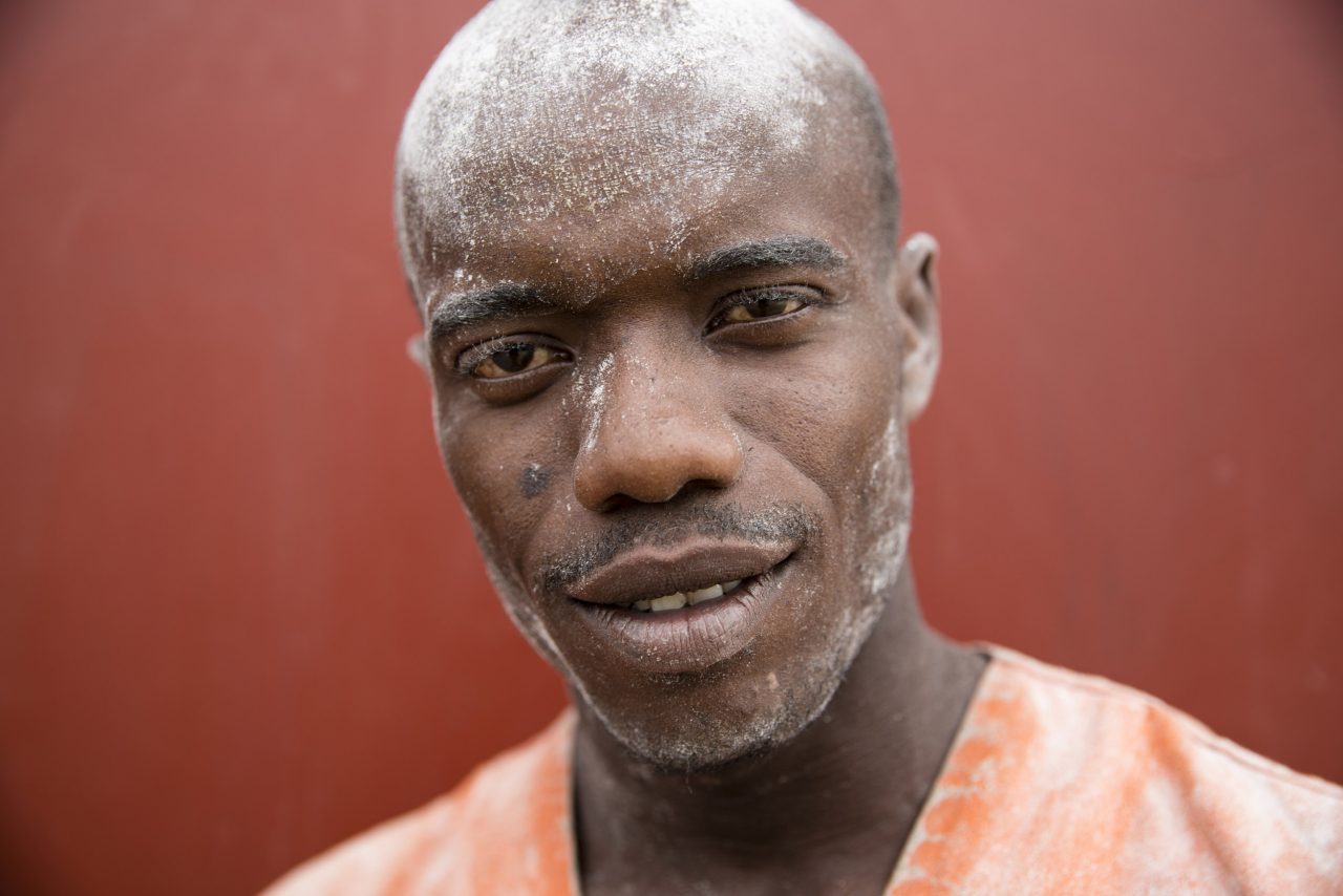 Portrait coporate d'auteur en gros plan sur fond rouge d'un jeune journalier le visage recouvert de farine sur le port industriel de Brazzaville au Congo, Il fixe l'objectif